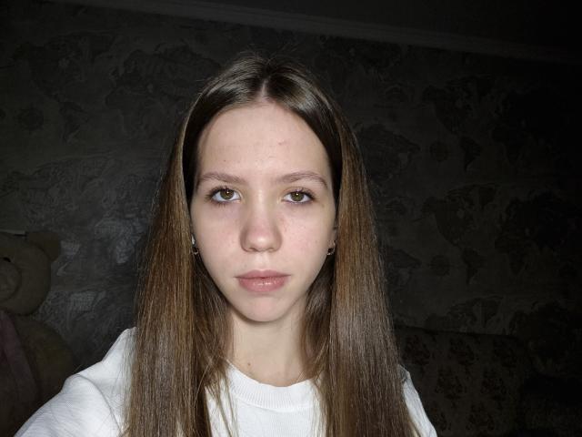 KarenPortal ha lunghi capelli castani, pelle chiara, piccoli seni e occhi marroni, indossa una camicia bianca e ha un'espressione seria in una stanza poco illuminata..