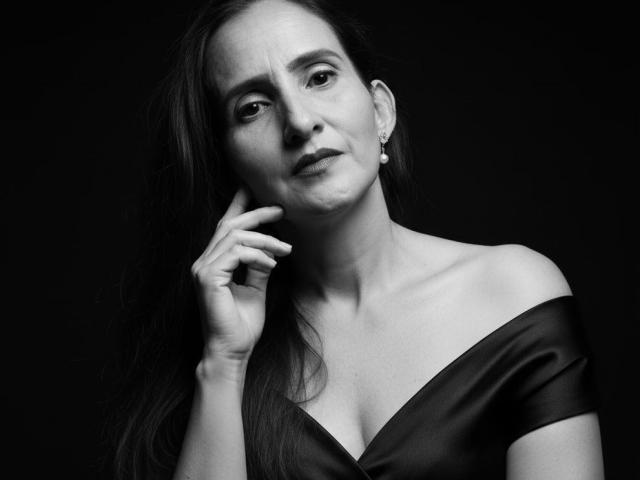 Black-and-white photo of RubiAnderson with long dark hair, off-shoulder satin top, small earrings, hand near chin, looking slightly confused, with visible collarbones and subtle smile.