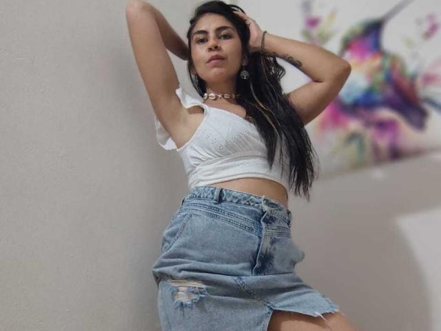 SweetShanny, com cabelos pretos longos, uma blusa cropped branca e jeans azuis rasgados, em pé contra uma parede, com os braços levantados, com uma expressão confiante, usando um colar, com um sorriso levemente convencido..