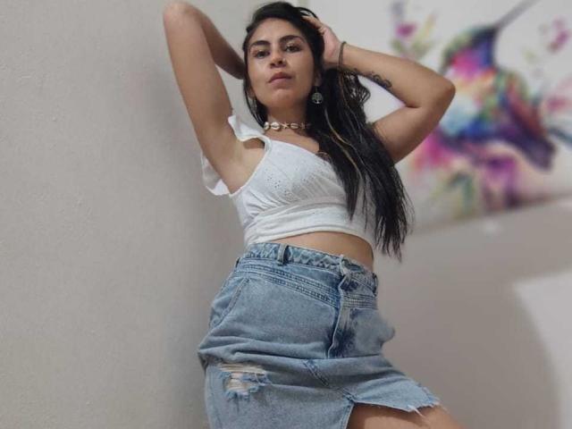 SweetShanny, con cabello largo y negro, un top blanco corto y una falda vaquera desgarrada de cintura alta, con los brazos levantados, luciendo segura de sí misma, usando un collar y una pulsera..