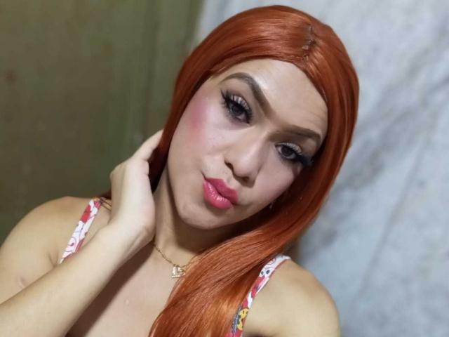 NathaliaLooes, avec ses cheveux roux, son rouge à lèvres rose et son eye-liner prononcé, se touche le cou, porte un haut fleuri, montre ses seins, sur une photo de profil prise avec une légère inclinaison..