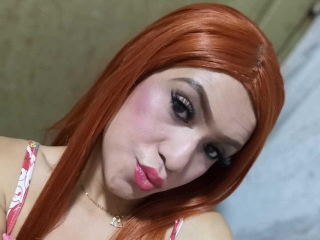 NathaliaLooes tiene el pelo rojo, labios rosados y delineador de ojos negro. Está haciendo una cara tonta, lleva una blusa rosa con estampado floral y tiene un collar dorado..