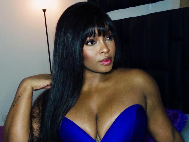 Nicoleth69, con cabello negro largo, vestido azul sin tirantes y busto grande, mirando a la cámara con labios rosados, brazo derecho levantado, piel oscura y tatuaje en el brazo izquierdo..