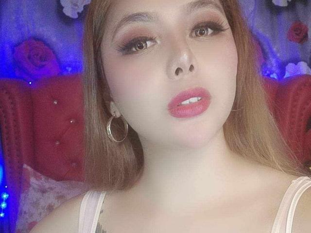 TikolyAma with pale skin, pink lips, and long brown hair, wearing a pink top and hoop earrings, looking at the camera with slightly parted pink lips.