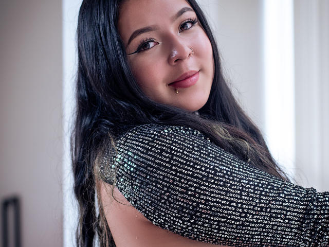 BarbaraHoneyy with long black hair, subtle makeup, nose ring, and septum piercing, wearing a sparkly black top, smiling gently at the camera. Her tits are partially visible.