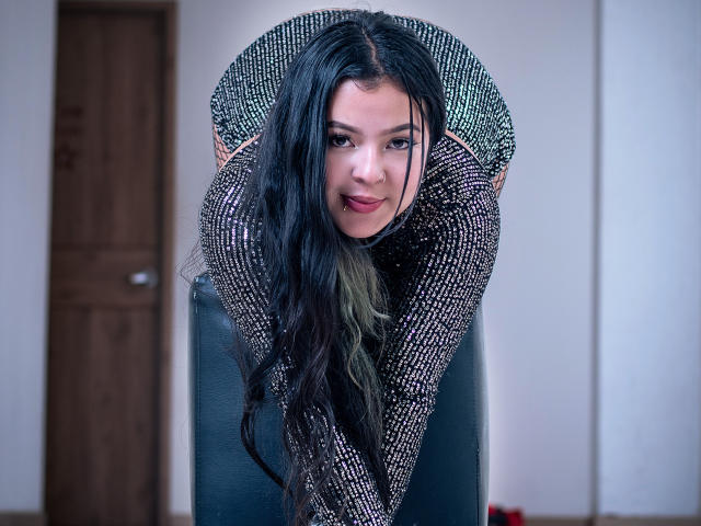 BarbaraHoneyy bent over, wearing a sparkly black long-sleeve top, with long black hair, green-tinted ends, and a playful smile, showing her ass and looking at the camera.