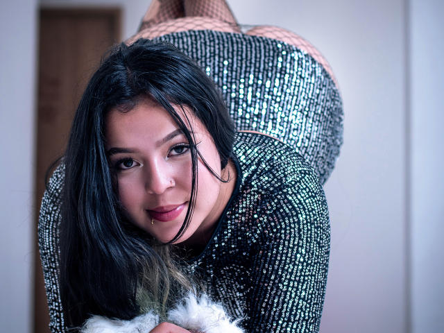 BarbaraHoneyy with black hair, nose ring, and septum piercing, bent over, wearing a sparkly black dress, fishnet stockings, and holding a furry white accessory, smiling at the camera.