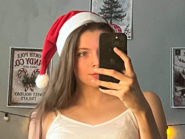 JennellMatacale with long brown hair, wearing a red Santa hat, white tank top, holding a black phone, taking a mirror selfie, showing her tits.