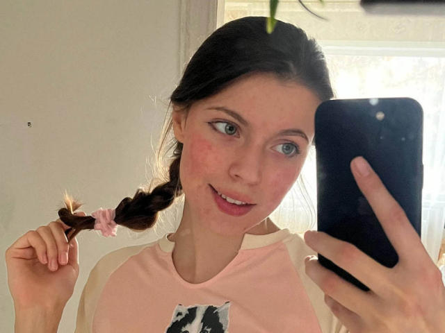 JennellMatacale with a braid in her right hand, holding a black phone, smiling with pink cheeks, wearing a pink and white shirt, in a selfie.