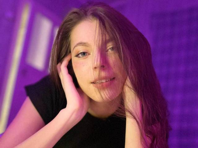 JennellMatacale with long brown hair, black shirt, hands on cheeks, purple lighting, slight smile, looking at camera, pinkish light in eyes, relaxed, soft light on face.