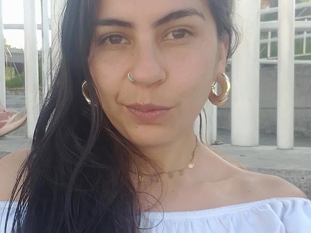SweetShanny hat langes, schwarzes Haar, braune Augen, einen Nasenring, goldene Ohrringe im Stecker-Design und eine kleine Halskette. Sie trägt ein weißes Top mit einem Schulterauschnitt, hat geschwollene Lippen und ein leichtes Lächeln..