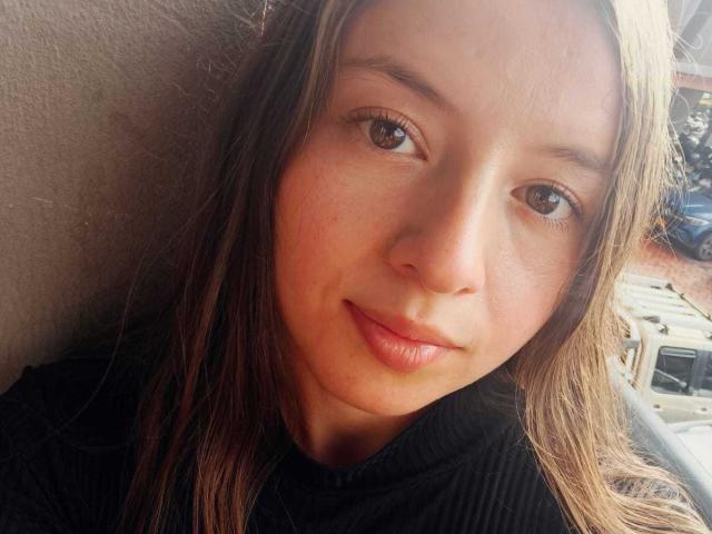 VioletaNova hat eine hellbraune Haut, braune Augen und langes, braunes Haar. Sie lächelt sanft mit rosafarbenen Lippen, trägt ein schwarzes, geripptes Oberteil und blickt direkt in die Kamera..