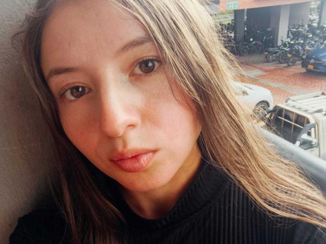 VioletaNova's face, with light brown eyes and slightly parted pink lips, in a black ribbed turtleneck, leaning against a gray wall. Blonde hair, fair skin.