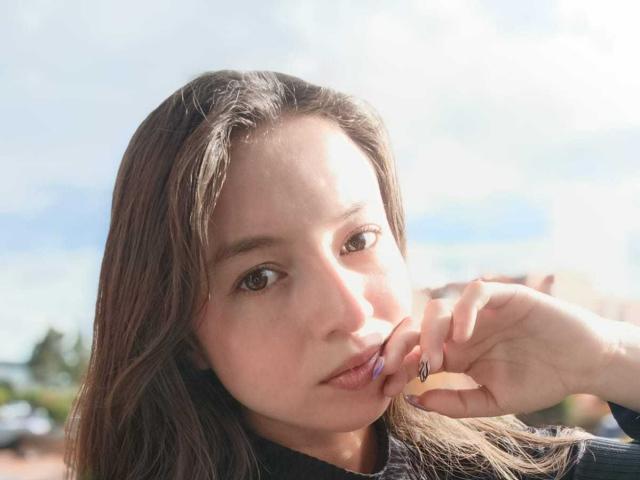 El rostro de VioletaNova, con cabello largo y castaño, ojos marrones y piel clara, mordiéndose el dedo con esmalte de uñas morado, vistiendo una blusa negra, bajo la luz brillante del sol..