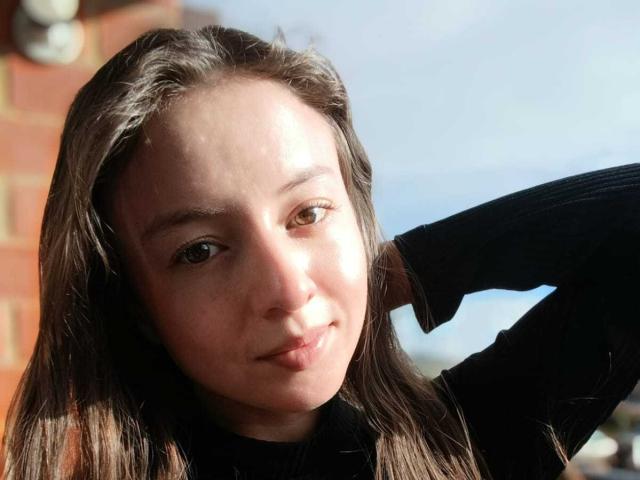 VioletaNova with puffy ass-adjacent face, brown eyes, black turtleneck, long brown hair, hand behind head, slight smile, sunlight on face, brick wall in background, clear blue sky.