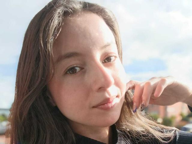 VioletaNova, avec une peau brun clair, des yeux marron et de longs cheveux bruns, touchant sa joue, souriant légèrement, portant un haut noir et arborant un petit anneau de nez..