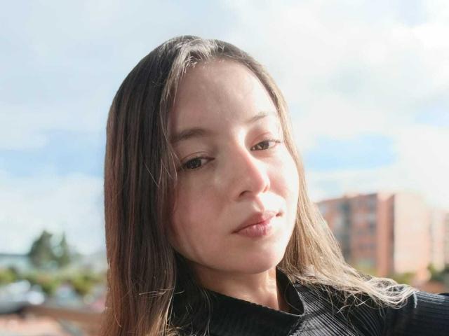 VioletaNova has straight brown hair, light skin, and is wearing a black shirt, with a subtle pout and a slight smile, in a bright, sunny outdoor setting.