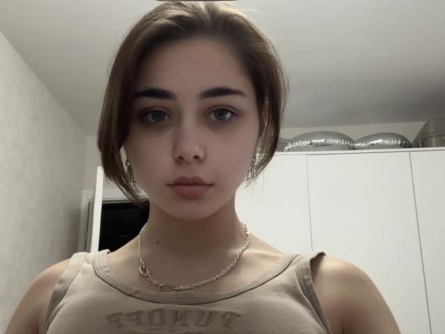 Janina Vidrio hat eine helle Haut, braune Augen und kurze, braune Haare. Sie trägt ein beiges Tanktop und eine Kette, und hat weiße Kopfhörer im Ohr..