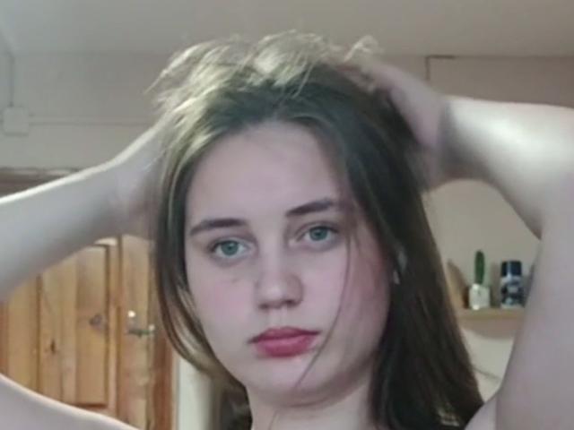 Alysia Finkelstein, com as mãos no cabelo castanho, olhos azuis, lábios rosados e pele clara, olhando para a câmera, sem roupas visíveis, em um quarto com pouca luz..
