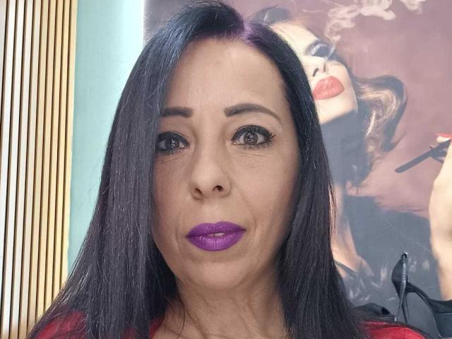 LauraSin with purple lipstick, black straight hair, and red top, looking serious with medium-sized tits, brown eyes, and subtle makeup. Background has a purple, smoky-eyed portrait.