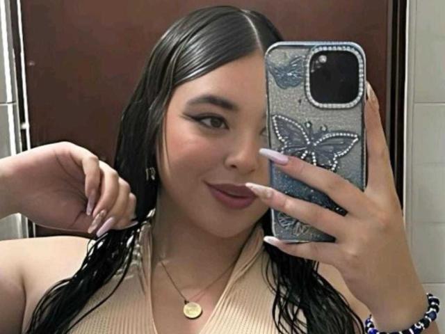 BaGiolett heeft nat zwart haar, draagt een beige top, houdt een telefoon vast met een hoesje met een vlinder erop, glimlacht, laat lange nagels zien en draagt een gouden ketting..
