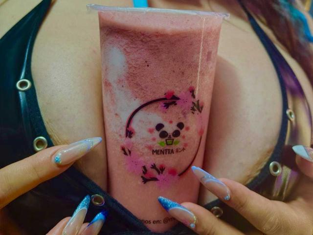 BaGiolett's handen houden een roze bevroren drankje vast, met een schattig beren gezicht en een roze bloemendesign. Haar lange, glinsterende blauwe nagels en zwarte top met metalen studs vormen een raam om het drankje..