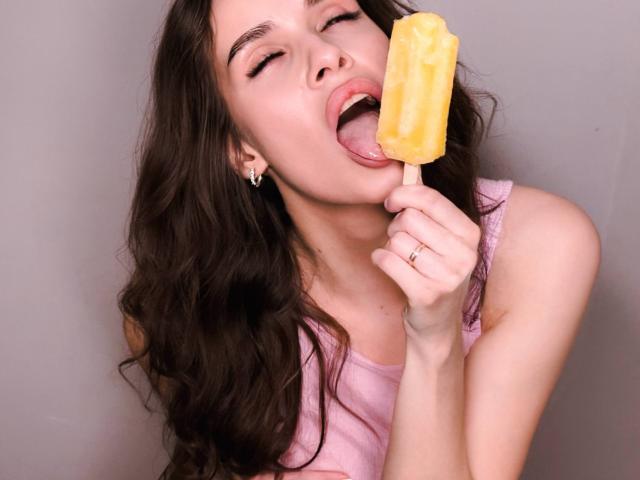 CandyCarrie with long brown hair, eyes closed, tongue out, licking a yellow popsicle, wearing a pink tank top, holding the stick with her right hand, ring on her finger.