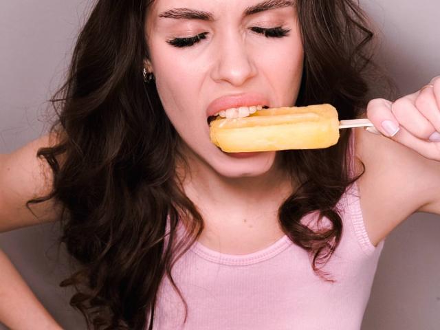 CandyCarrie, con cabello largo y castaño, camiseta rosa, comiendo una paleta amarilla con palo, boca abierta, mostrando los dientes, mirando hacia abajo, sosteniendo el palo con la mano derecha..