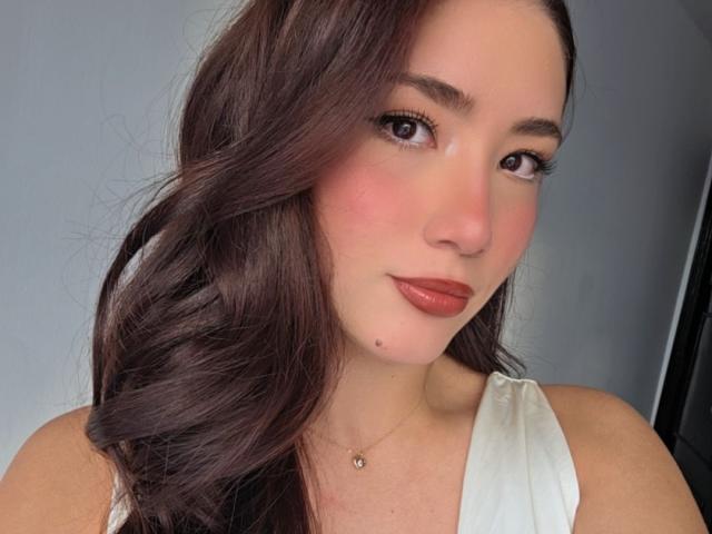 Stardustt has dark brown wavy hair, pink blushed cheeks, and glossy brown eyes. She's wearing a white sleeveless top and a delicate necklace. Her lips are slightly parted.