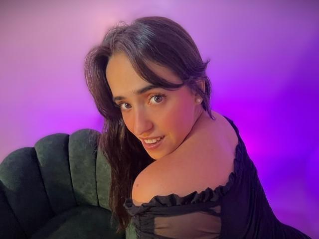 AlysonFox with long brown hair, black off-shoulder top, and purple-blue lighting, smiling over her shoulder, showing her bare shoulder and green velvet chair.