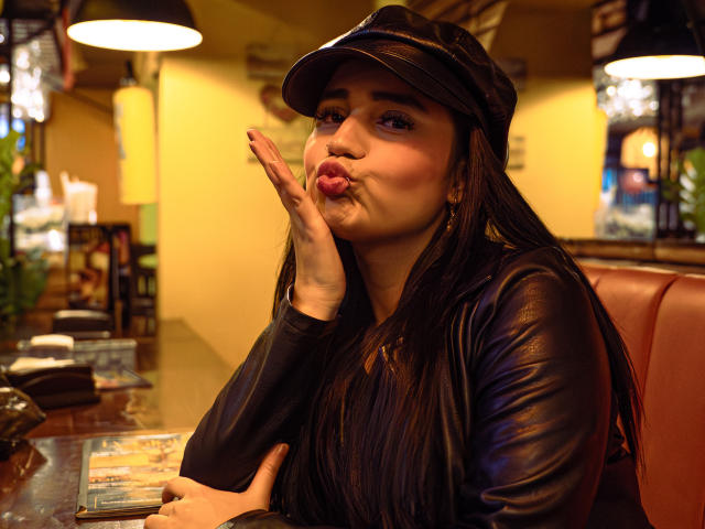 JulietaDupont with pouty lips, dark leather cap, and long brown hair, making a kissing face, hand up, wearing a black leather jacket, sitting at a bar.