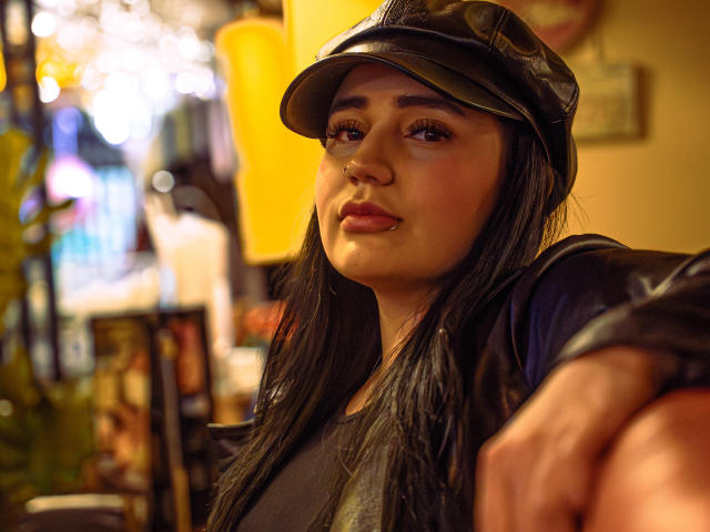 JulietaDupont with dark hair, wearing a black leather cap and jacket, has a cute nose ring and pouty lips, her brown eyes looking to the side, in a cozy, lit-up spot.