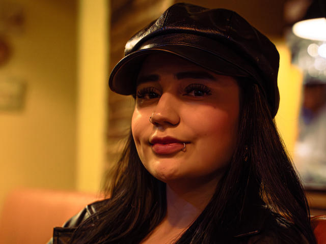 JulietaDupont with black leather cap, long dark hair, nose ring, lip piercing, and pink lipstick, wearing black leather jacket, smiling softly, with warm yellow lighting.