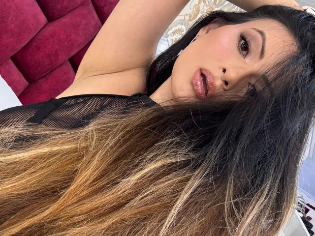 HannaRobertss with long, ombre hair, black mesh top, and glossy lips, lying on a red, tufted bed, arms above head, looking at camera with sultry expression.