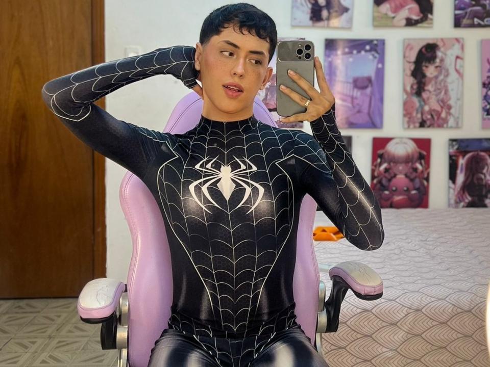 SweetCamellia, con cabello negro rizado, vestida con un traje negro de Spider-Man, sentada en una silla rosa, tomando un selfie con un iPhone, con las manos detrás de la cabeza, y los labios ligeramente entreabiertos..