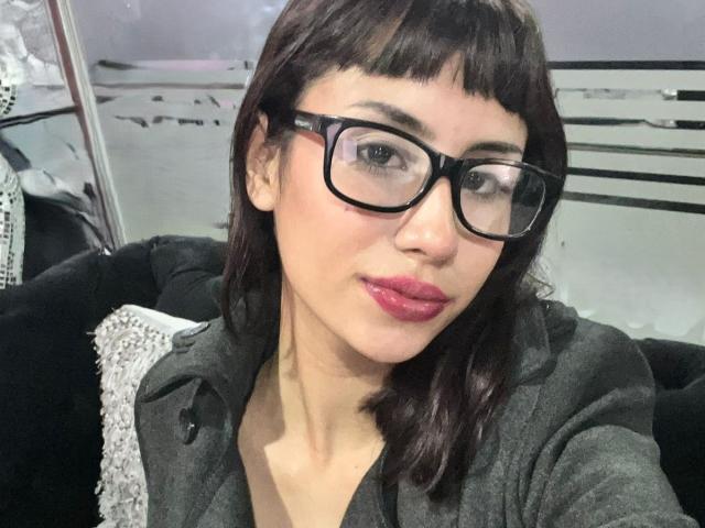EmilyDubois, com cabelo castanho escuro, óculos pretos, batom rosa e moletom cinza, olhando para a câmera com um leve sorriso. Seus seios estão ligeiramente visíveis sob o moletom..