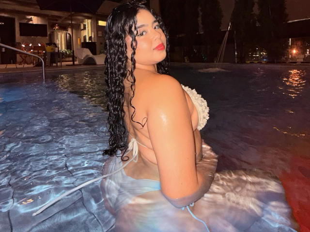 AprielKeith with wet, curly black hair, red lipstick, and a white floral bikini, showing her big ass and shiny tits in a nighttime pool, looking back over her shoulder.