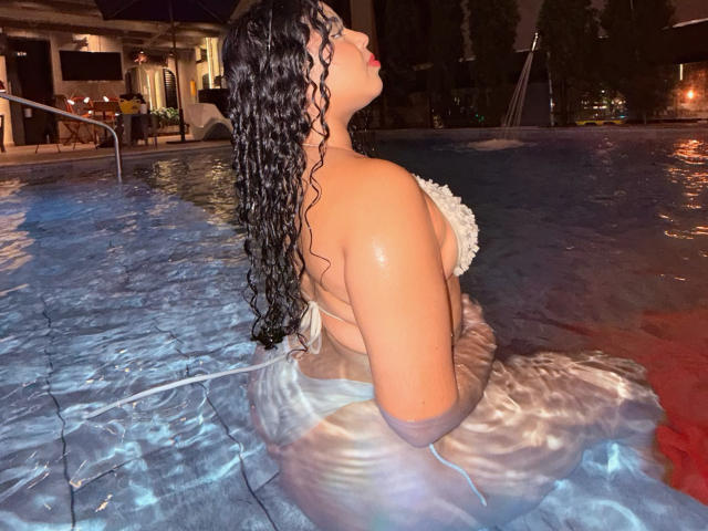 AprielKeith with wet curly black hair, large tits, and a white ruffled bikini top, standing in a pool at night, looking upward with red lipstick.