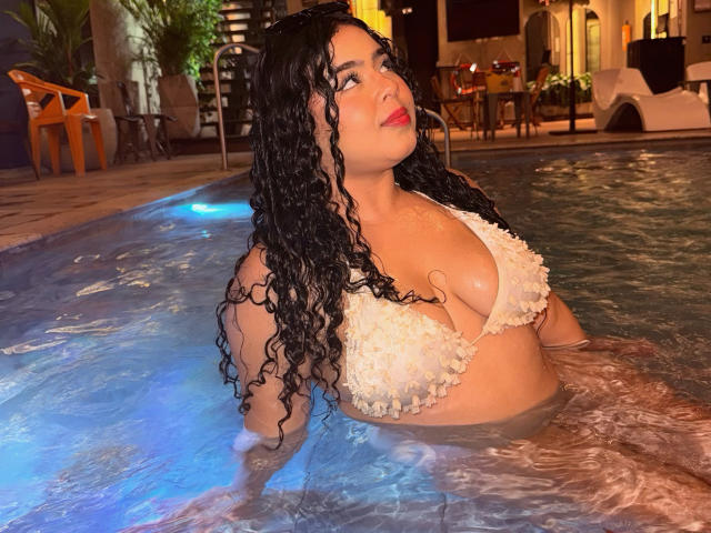 AprielKeith with curly black hair, wet, in a pool, wearing a frilly white bikini, showing large tits, looking up, red lipstick, night, water reflecting light.