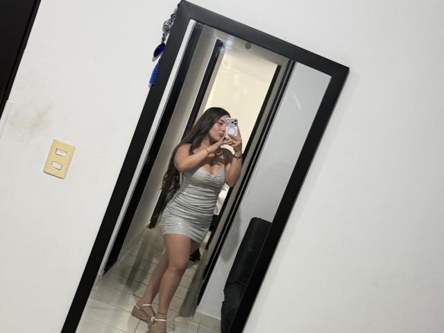 LorensDouce in a tight silver dress, holding a phone, taking a mirror selfie, showing off her ass and long brown hair, wearing strappy heels, in a white-walled hallway.