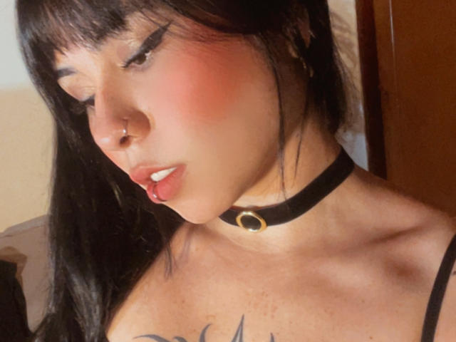 GaiaJoness, con mejillas rosadas, un collarín negro, un piercing en la nariz y un piercing en el labio, mirando hacia abajo, con cabello oscuro, un tatuaje en el pecho y labios entreabiertos. Sus pechos son ligeramente visibles..