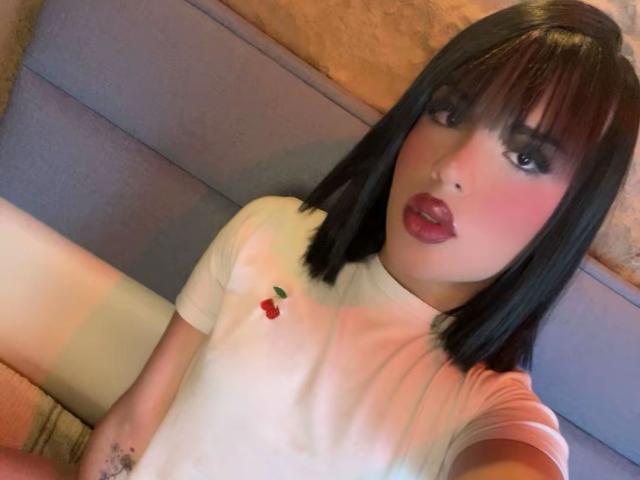 GlossyyBratz with black bob, pink blush, red lipstick, white shirt with cherry embroidery, sitting on gray sofa, right arm visible, subtle tattoo on wrist, selfie.
