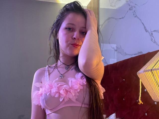 GraceRedMiller, con i capelli castani bagnati, reggiseno rosa con fiori e piercing al naso, che si tocca la testa, guarda la fotocamera, mostrando piccoli seni, in una stanza poco illuminata, indossando una collana..