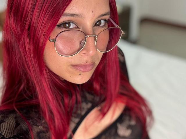 CherryMist with red hair, wearing glasses, black lace top, and a nose piercing, showing her pouty lips and brown eyes. Her tits are subtly visible through the lace.