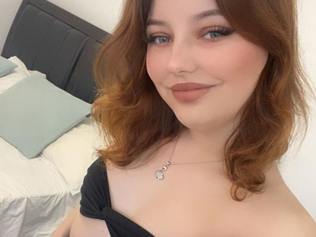 LilSoft has wavy brown hair, blue eyes, and a cute smile. She's wearing a black strap top, showing her boobs, with a silver necklace and subtle makeup.