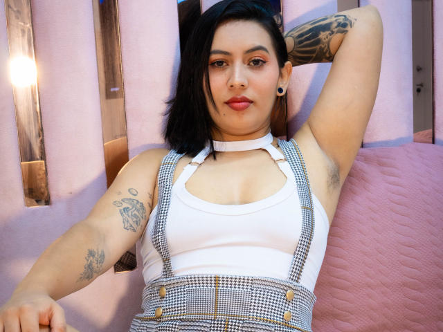 Amy Ross, com cabelo preto, vestindo uma regata branca e suspensórios xadrez, tem tatuagens em ambos os braços, mostrando suas axilas, e está sentada encostada em um cobertor rosa..