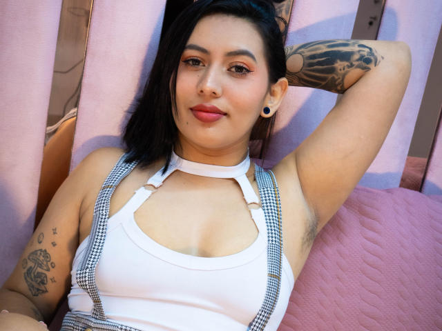 AmyRoss with black hair, tattoos, and black ear gauges, wearing a white tank top with checkered suspenders, arm raised, pink quilt, smiling, showing her tits.