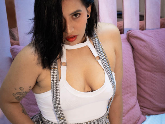 AmyRoss with black hair, red lipstick, white tank top, houndstooth suspenders, showing her tits, tattoo on left arm, sitting on pink pillows, looking at camera.