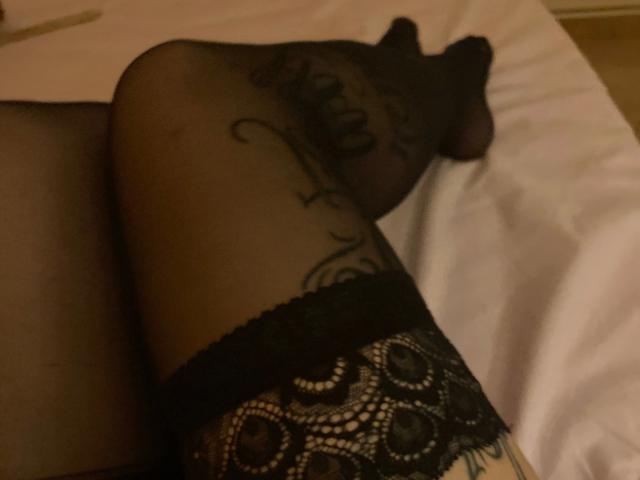 MelissaStrip-hot's black lace thigh-highs, showing a tattoo on her right thigh, with her legs crossed on a white bed. Her bare, smooth, tan-skinned ass is partially visible.