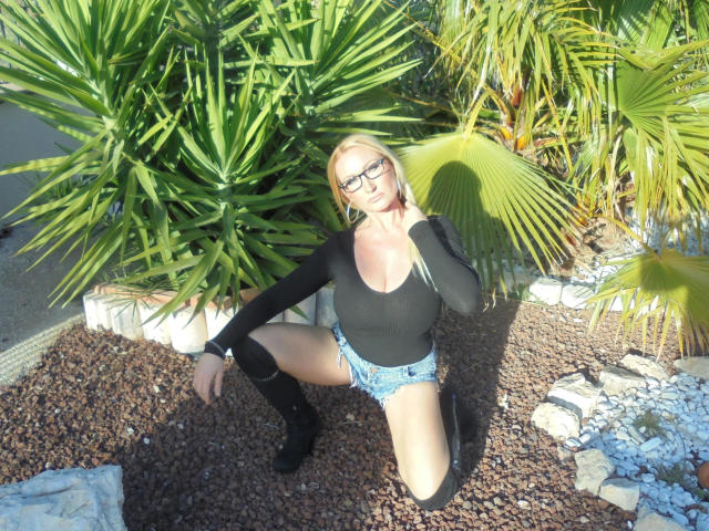 MelissaStrip-hot with blonde hair, black glasses, black long-sleeve top, blue denim shorts, black knee-high boots, crouching in gravel, palm plants behind, sunlight, shadow.